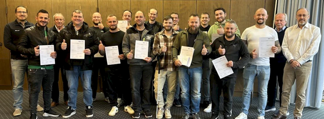 Group of 18 men in a casual look, 15 of whom are holding the thumbs up and a certificate in their hand because they have successfully completed further training to become foremen in furnace and chimney construction