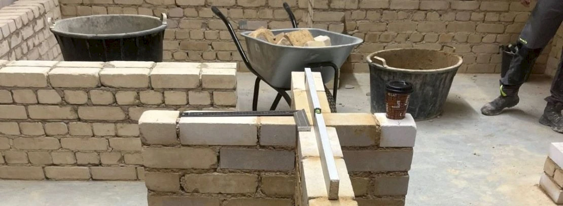 Photo with a wall cross model on it there is a square and a spirit level for measuring. In the background there is a wheelbarrow, more walls and a mortar bucket